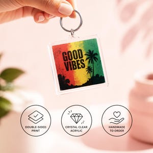 Pode incluir: Um porta-chaves quadrado em acrílico com um fundo degradé vermelho, amarelo e verde, silhuetas pretas de palmeiras e o texto GOOD VIBES impresso a preto. O porta-chaves inclui um anel de metal prateado para fixação.
