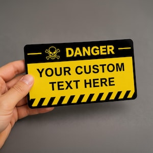 May include: A rectangular sign with a black and yellow color scheme. The top section features a skull and crossbones icon next to the word DANGER. The center displays the text YOUR CUSTOM TEXT HERE in black, and the bottom has a striped border.