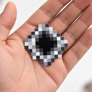 Puede incluir: Un pin de esmalte en forma de diamante con un diseño pixelado. El centro es un cuadrado negro sólido rodeado por capas de cuadrados en varios tonos de gris y blanco, creando un efecto de degradado que imita un gráfico digital de baja resolución.