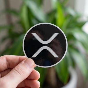 Op de afbeelding: Een ronde, zwart-witte sticker met het Ripple cryptocurrency logo. Het logo is een gestileerde witte 'X' op een zwarte achtergrond. De sticker wordt voor een vage groene plant gehouden.