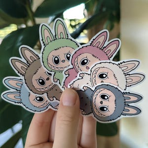 May include: A collection of colorful stickers featuring a cartoon character in a bunny costume. The stickers have a white border and are in shades of green, pink, gray, and brown. The character has large eyes and a toothy grin.