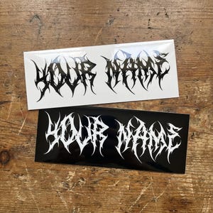 Puede incluir: Dos pegatinas rectangulares de vinilo con el texto YOUR NAME en una fuente de estilo gótico afilada y dentada. Una pegatina tiene texto negro sobre fondo blanco y la otra texto blanco sobre fondo negro, ambas con un acabado brillante.