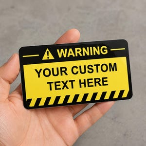Può includere: Un cartello di avvertimento rettangolare con un bordo superiore nero contenente la parola WARNING e un'icona con punto esclamativo. Il centro è giallo con il testo YOUR CUSTOM TEXT HERE in nero. La parte inferiore presenta strisce diagonali nere e gialle.