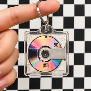 May include: A square, clear acrylic keychain featuring a miniature replica of a MiniDisc. The disc has a holographic, iridescent surface with the number 80 printed in black, a small MiniDisc logo, and a silver-colored metal shutter detail.
