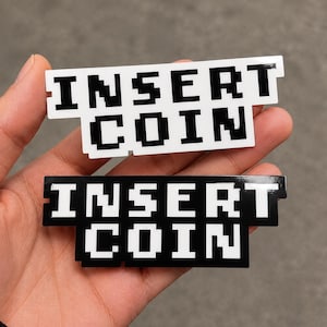 Peut inclure: Deux autocollants rectangulaires avec le texte pixélisé INSERT COIN. Un autocollant a un fond blanc avec des lettres noires, et l'autre a un fond noir avec des lettres blanches. Les deux autocollants ont une finition brillante.