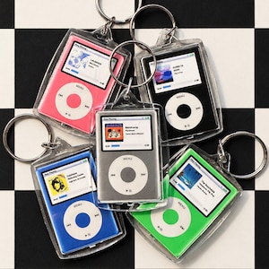 Puede incluir: Cinco llaveros de acrílico con forma de reproductores de música digitales en rosa, negro, plata, azul y verde. Cada uno cuenta con una pantalla que muestra una interfaz de reproducción con detalles de pista y una rueda de control circular.