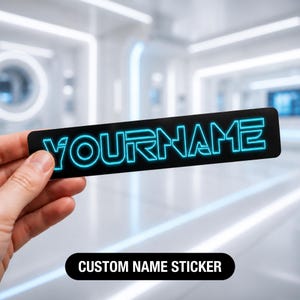 Könnte beinhalten: Ein rechteckiger schwarzer Aufkleber mit dem Wort YOURNAME in einer leuchtenden, cyanblauen Neon-Schriftart. Die Buchstaben haben einen Glüheffekt vor dem dunklen Hintergrund, was für einen hohen Kontrast bei diesem Namensaufkleber sorgt.