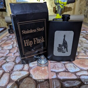 May include: A black stainless steel hip flask with a Wild Turkey Bourbon logo. The flask is sitting on a stone-patterned surface. The flask comes in a black box with the words "Stainless Steel Hip Flask" printed on it.