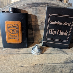 May include: A black stainless steel hip flask with a brown leather patch featuring the Jack Daniel's logo. The flask is sitting on a wooden surface.