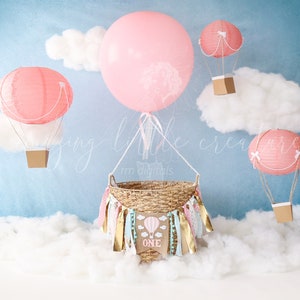 May include: A pink and white hot air balloon themed baby shower backdrop with a basket, clouds, and a banner that says "One".