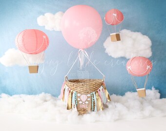 May include: A pink and white hot air balloon themed baby shower backdrop with a basket, clouds, and a banner that says "One".