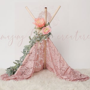 Könnte beinhalten: Ein rosa Spitzen-Tipi mit Holzstangen und einem Blumengirlanden am oberen Ende. Das Tipi steht auf einer weißen, flauschigen Oberfläche. Der Text "amazing little creatures" ist in rosa Schrift auf der weißen Wand hinter dem Tipi geschrieben.