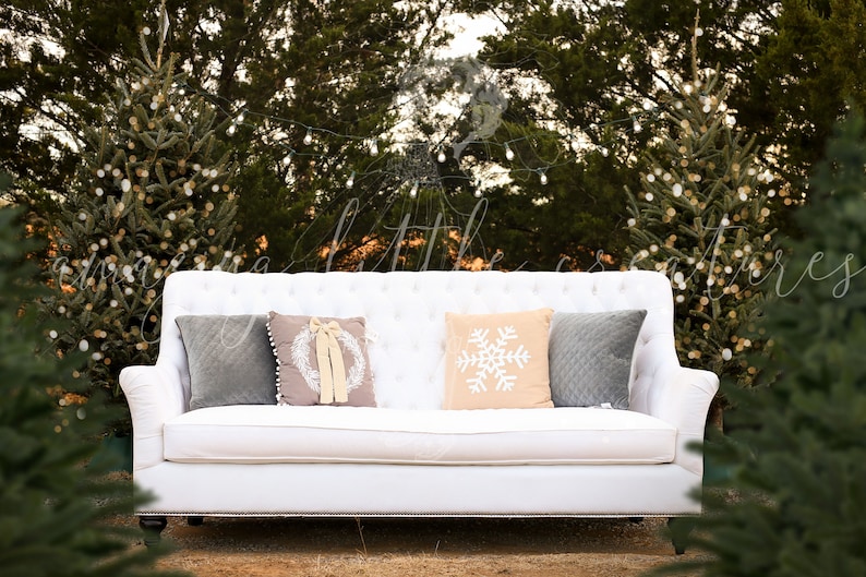 May include: A white tufted sofa with four pillows, two gray, one with a wreath design, and one with a snowflake design, sits in front of a backdrop of evergreen trees with white lights.