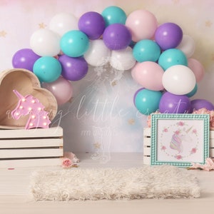 May include: A unicorn themed birthday party backdrop with a white, pink, purple and teal balloon arch, a wooden heart-shaped shelf with a pink unicorn light, a white wooden crate, a white rug, and a teal frame with a unicorn illustration.
