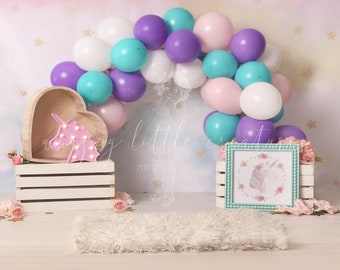 May include: A unicorn themed birthday party backdrop with a white, pink, purple and teal balloon arch, a wooden heart-shaped shelf with a pink unicorn light, a white wooden crate, a white rug, and a teal frame with a unicorn illustration.