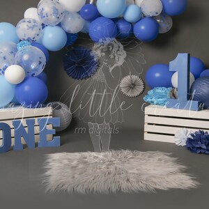 May include: A blue and white balloon arch with a blue number one and the word "ONE" in blue letters. The balloons are arranged in a semi-circle shape and are decorated with confetti. There are also blue and white paper flowers and a white crate with a blue and white rug in front of it.