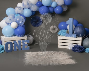May include: A blue and white balloon arch with a blue number one and the word "ONE" in blue letters. The balloons are arranged in a semi-circle shape and are decorated with confetti. There are also blue and white paper flowers and a white crate with a blue and white rug in front of it.