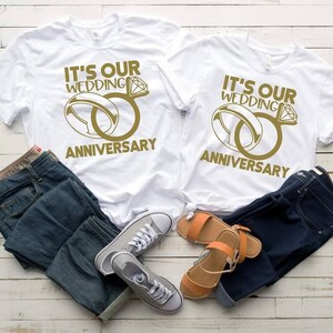 May include: Two white t-shirts with the text "It's our wedding anniversary" and a gold ring design. The shirts are laid out on a white wooden surface with blue jeans and sandals.