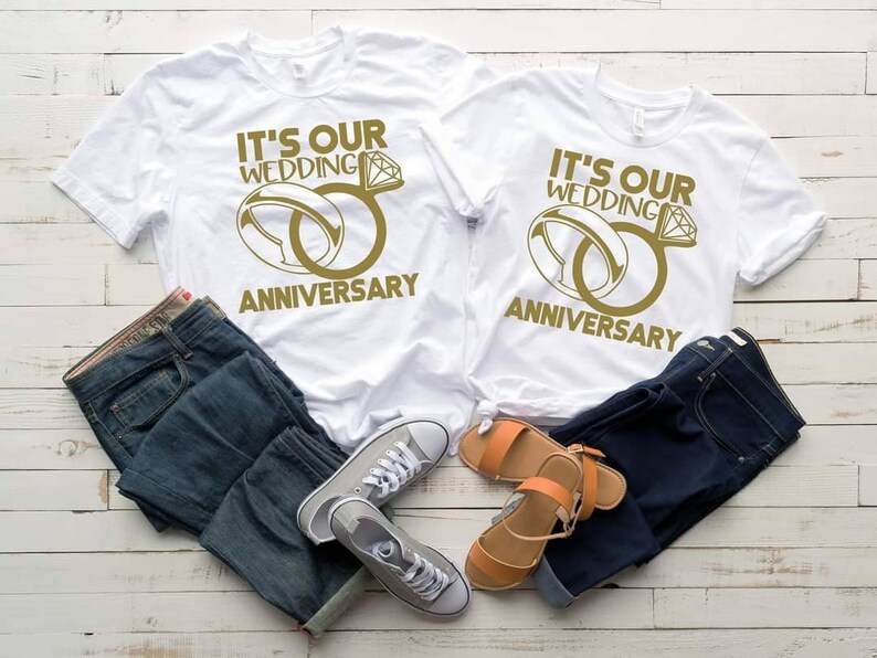May include: Two white t-shirts with the text "It's our wedding anniversary" and a gold ring design. The shirts are laid out on a white wooden surface with blue jeans and sandals.