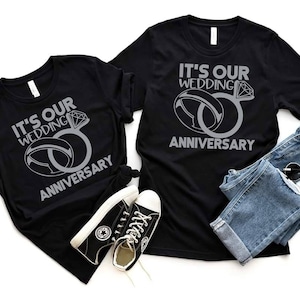May include: Two black t-shirts with the text "It's our wedding anniversary" and a silver ring design.
