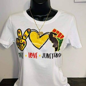 Pode incluir: Camiseta branca com um design gr&aacute;fico com um sinal de paz, um cora&ccedil;&atilde;o amarelo e um punho erguido. As palavras "PEACE LOVE JUNETEENTH" s&atilde;o impressas abaixo. Um colar de ouro com um pingente tamb&eacute;m &eacute; vis&iacute;vel.