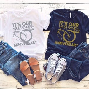 May include: Two t-shirts with the text "It's our wedding anniversary" and a graphic of two interlocking rings. One shirt is white with silver rings and the other is black with gold rings.