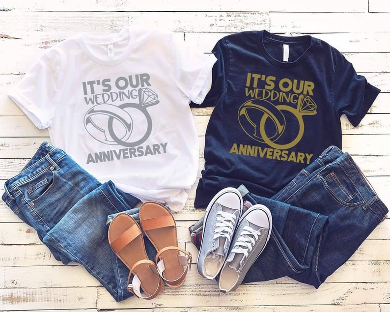 May include: Two t-shirts with the text "It's our wedding anniversary" and a graphic of two interlocking rings. One shirt is white with silver rings and the other is black with gold rings.