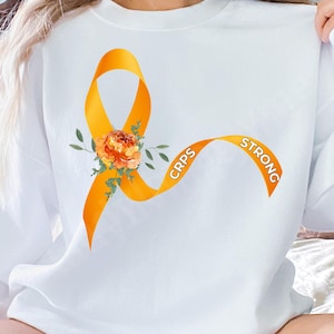 Könnte beinhalten: Weißes Sweatshirt mit einem orangefarbenen Band mit der Aufschrift "CRPS STRONG" und einem Blumendesign. Das Band ist ein Symbol für das Bewusstsein für das komplexe regionale Schmerzsyndrom.