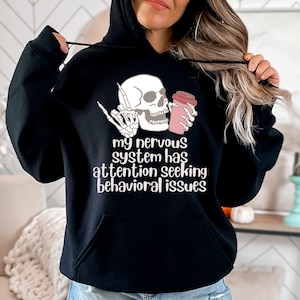 May include: Black hoodie with a white skull holding a pink coffee cup. The text on the hoodie reads "My nervous system has attention seeking behavioral issues."