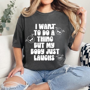 May include: Dark gray t-shirt with the text "I WANT TO DO A THING BUT MY BODY JUST LAUGHS" in white, along with three laughing face graphics. The shirt is worn with blue jeans.