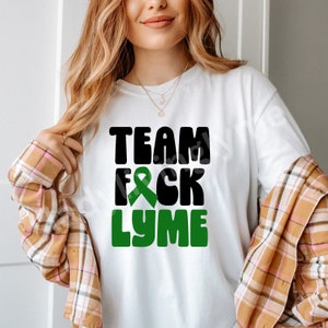 May include: White t-shirt with black text that reads "TEAM FCK LYME" with a green ribbon in the middle of the text.