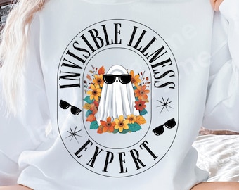 May include: White sweatshirt with a graphic of a ghost in sunglasses, encircled by flowers. The text reads "INVISIBLE ILLNESS EXPERT" in a circular design, with additional sunglasses and starbursts.
