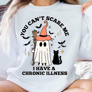 Puede incluir: Sudadera blanca con un diseño de Halloween. El diseño presenta un fantasma con un sombrero de bruja y una escoba, con el texto "YOU CAN'T SCARE ME I HAVE A CHRONIC ILLNESS". Incluye murciélagos, estrellas y un gato negro.