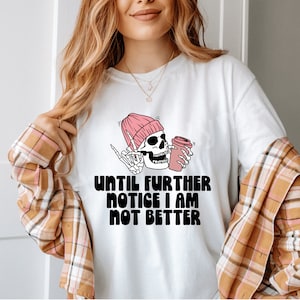 May include: White t-shirt with a graphic of a skeleton wearing a pink beanie and holding a coffee cup. The text "Until further notice I am not better" is printed below the graphic.