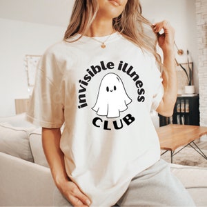 May include: White t-shirt with a black graphic of a ghost with the text "invisible illness club" around it.