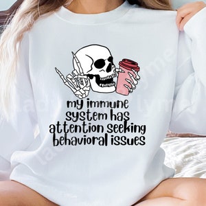 May include: White sweatshirt featuring a black and white illustration of a skeleton holding a pink coffee cup. The text "my immune system has attention seeking behavioural issues" is printed below the illustration.