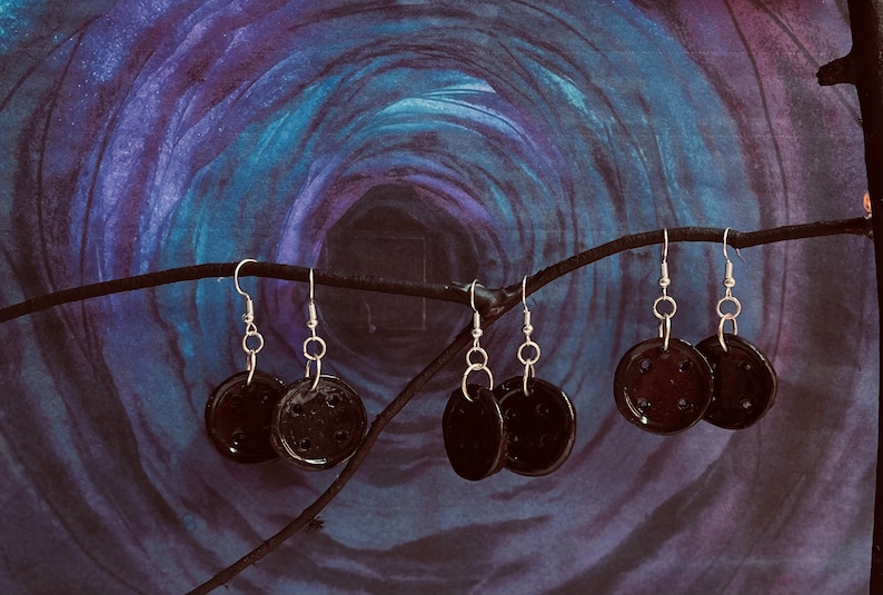 Puede incluir: Un conjunto de seis pendientes de cer&aacute;mica negra con ganchos plateados. Los pendientes son redondos y tienen una superficie texturizada. Est&aacute;n colgados de una rama sobre un fondo de acuarela azul y morado.