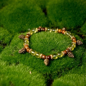 May include: A beaded bracelet with a nature theme. The bracelet features light yellow and brown beads, with small pinecone charms. The bracelet is set against a background of green moss.
