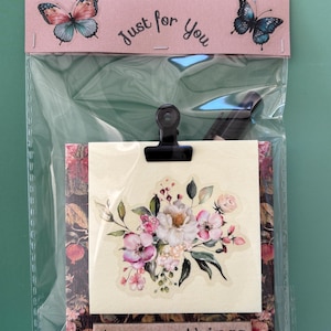 May include: A clear plastic bag containing a square note card with a floral illustration, held by a black metal clip. The card features a pink header with two butterflies and the text Just for You, and a bottom label with the text try new things.