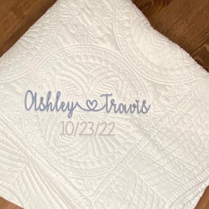 May include: A white quilted blanket featuring an intricate stitched pattern throughout. Embroidered in blue thread across the center is the text Ashley, a heart symbol, and Travis, with the date 10/23/22 stitched in blue thread below the names.