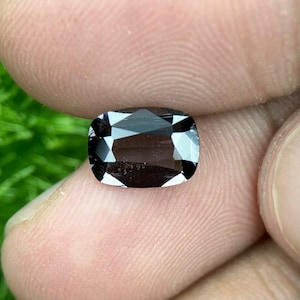 May include: A dark grey, cushion-cut gemstone with multiple facets. The gem's surface is polished, reflecting light and creating a sparkling effect. The stone is held between fingers, highlighting its shape and colour.