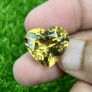 Puede incluir: Una gema facetada en forma de corazón con un tono amarillo verdoso. La gema se sostiene sobre un fondo de hierba verde. El corte de la piedra crea múltiples superficies reflectantes.