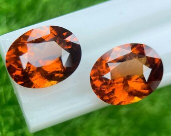 May include: Two oval-shaped orange gemstones with a faceted cut. The gemstones are set on a white surface.