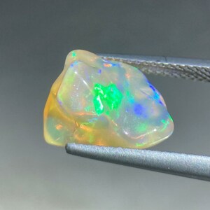 May include: A rough, white opal gemstone with green, blue, and orange flashes of colour. The opal is held in tweezers.