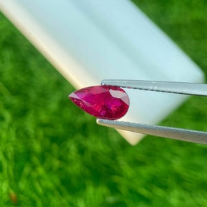 May include: A pear-shaped ruby gemstone held by tweezers. The ruby is a deep red color and has a high level of brilliance.