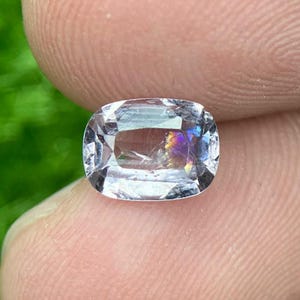 May include: A clear, rectangular gemstone with rounded corners. The gem has internal rainbow-coloured refractions. The stone is held between two fingers, with a blurred green background.