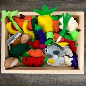 Op de afbeelding: Een houten kist gevuld met kleurrijke viltvoedingsmiddelen. Het assortiment omvat een ananas, bananen, wortelen, een watermeloen schijf, een vis en diverse andere groenten en fruit. De kist is gemaakt van lichtgekleurd hout.