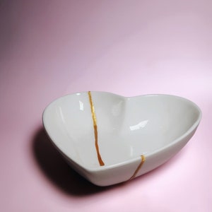 May include: White heart-shaped ceramic bowl with gold accents. The bowl is on a pink background.