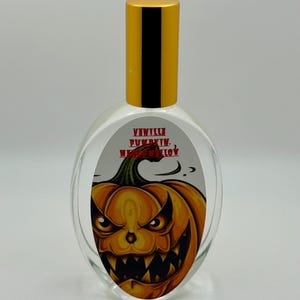 May include: Clear glass perfume bottle with a gold cap. The label features a menacing orange jack-o'-lantern with the words "Vanilla Pumpkin Marshmallow" in red. The bottle has an oval shape.