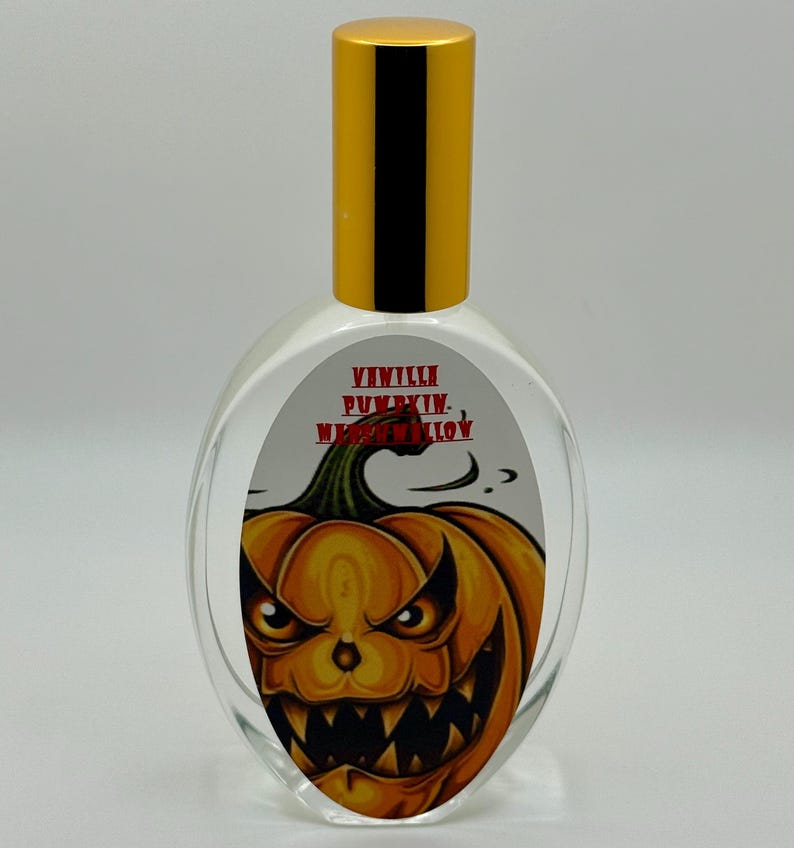 May include: Clear glass perfume bottle with a gold cap. The label features a menacing orange jack-o'-lantern with the words "Vanilla Pumpkin Marshmallow" in red. The bottle has an oval shape.